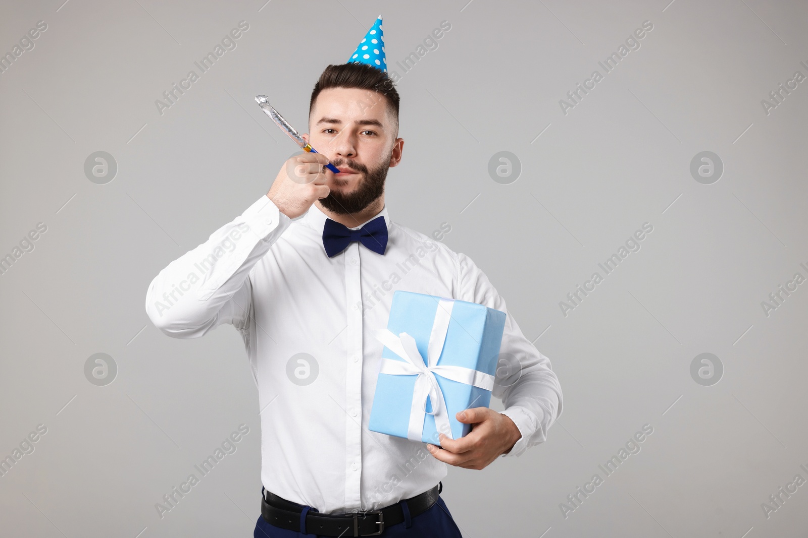 Happy Birthday. Handsome man with party hat, blower and gift box on grey background Photo of Happy Birthday. Handsome man with party hat, blower and gift box on grey background