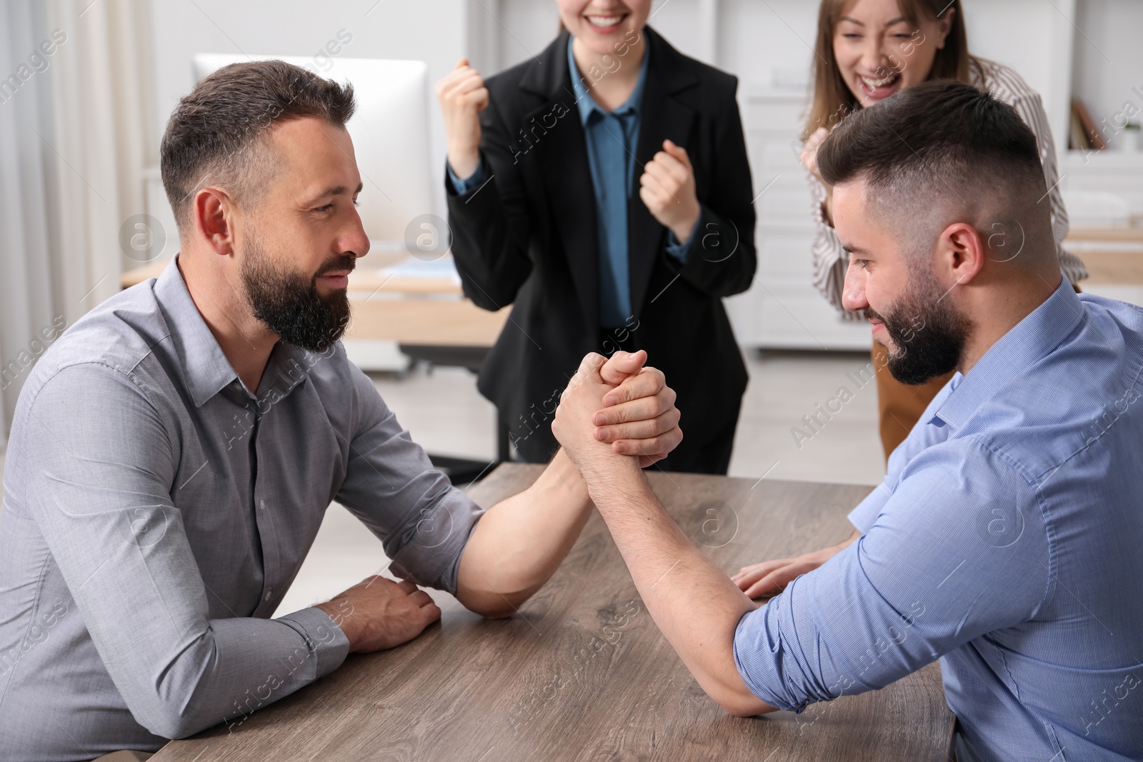 Businesspeople arm wrestling at wooden table in office Photo of Businesspeople arm wrestling at wooden table in office