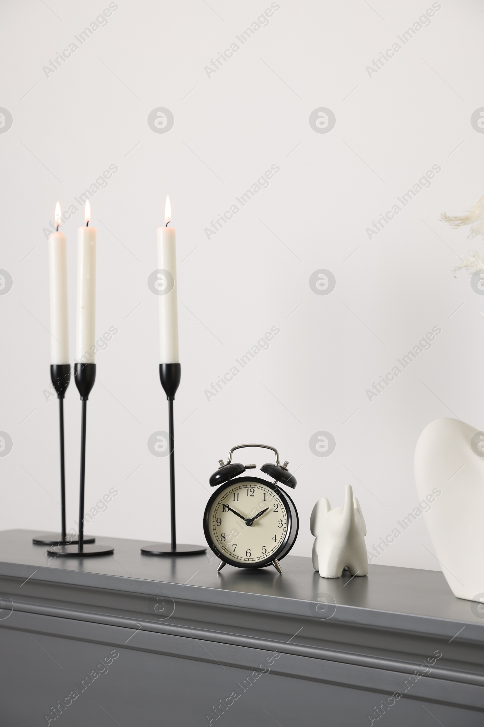 Stylish candlesticks with burning candles, other decor and alarm clock on fireplace near white wall indoors Photo of Stylish candlesticks with burning candles, other decor and alarm clock on fireplace near white wall indoors