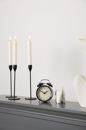 Photo of Stylish candlesticks with burning candles, other decor and alarm clock on fireplace near white wall indoors