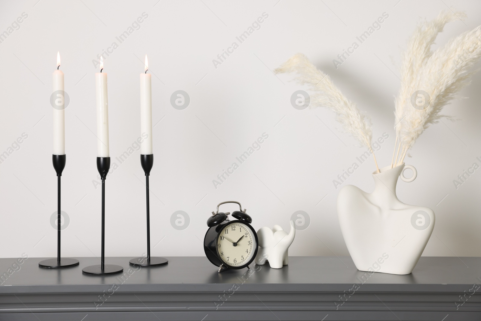 Stylish candlesticks with burning candles, other decor and alarm clock on fireplace near white wall indoors Photo of Stylish candlesticks with burning candles, other decor and alarm clock on fireplace near white wall indoors