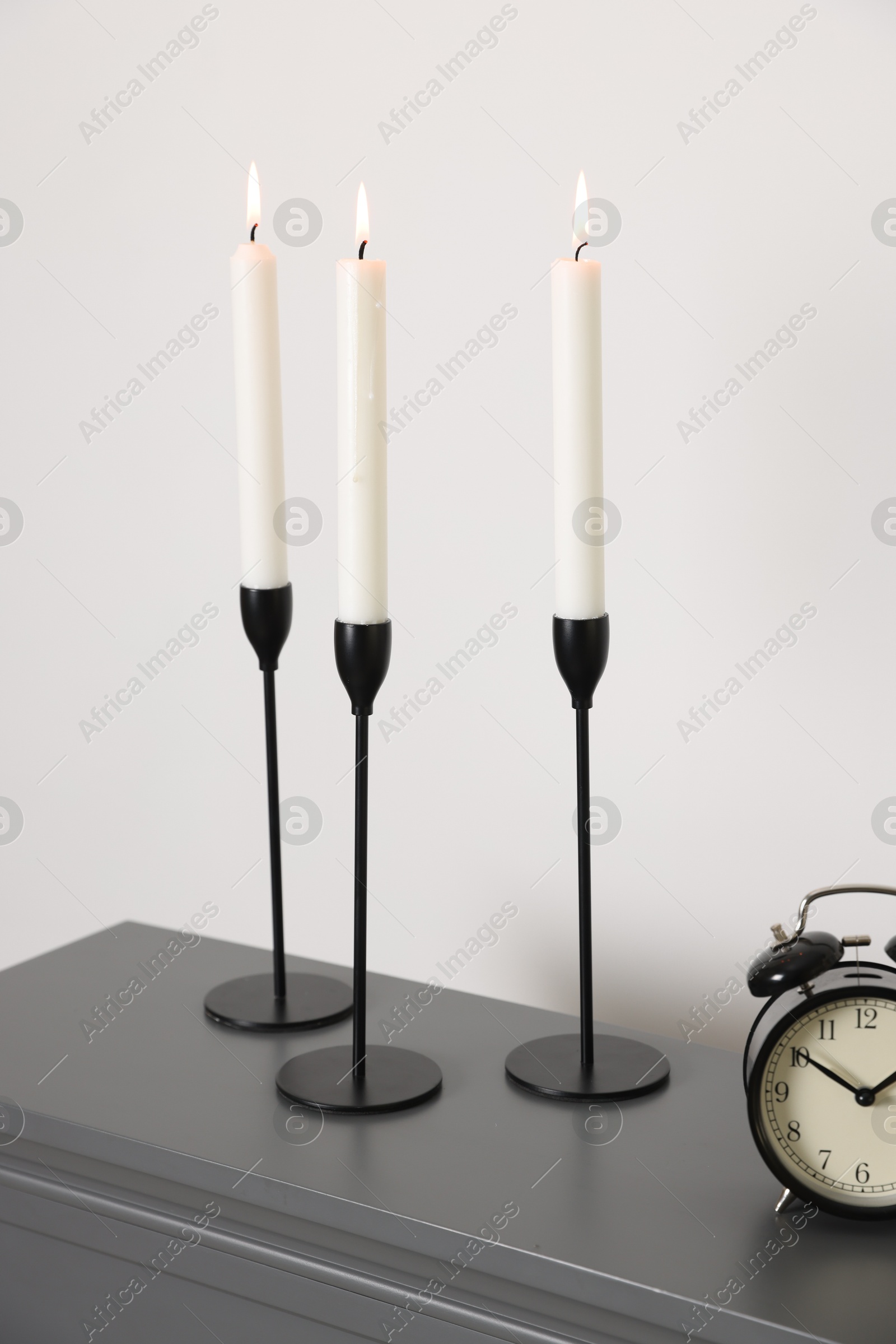Stylish candlesticks with burning candles and alarm clock on fireplace near white wall indoors Photo of Stylish candlesticks with burning candles and alarm clock on fireplace near white wall indoors