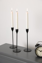 Photo of Stylish candlesticks with burning candles and alarm clock on fireplace near white wall indoors