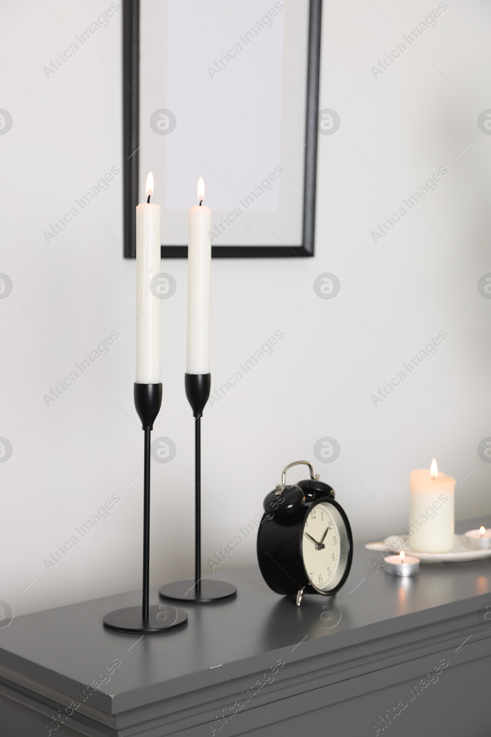 Stylish candlesticks with burning candles and alarm clock on fireplace near white wall indoors Photo of Stylish candlesticks with burning candles and alarm clock on fireplace near white wall indoors