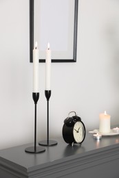 Photo of Stylish candlesticks with burning candles and alarm clock on fireplace near white wall indoors