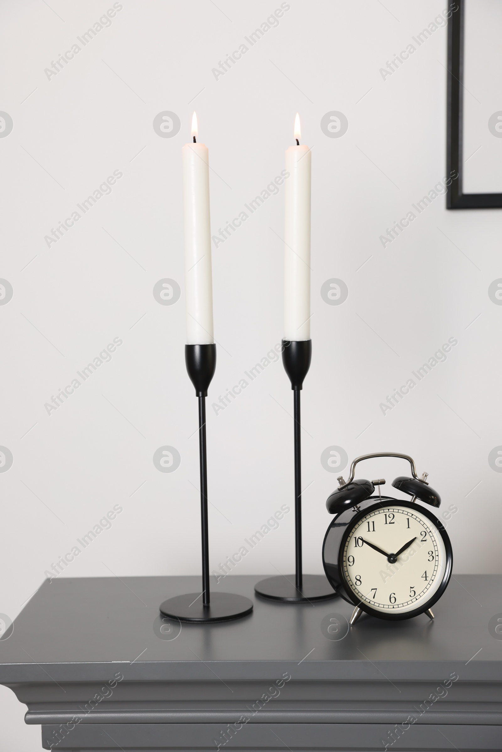 Stylish candlesticks with burning candles and alarm clock on fireplace near white wall indoors Photo of Stylish candlesticks with burning candles and alarm clock on fireplace near white wall indoors
