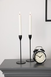 Photo of Stylish candlesticks with burning candles and alarm clock on fireplace near white wall indoors
