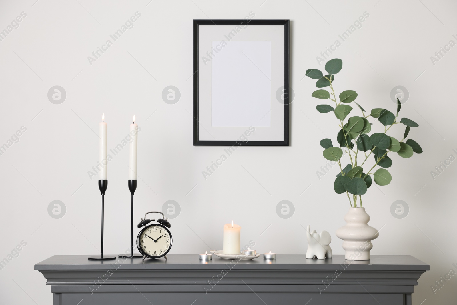 Photo of Stylish candlesticks with burning candles, other decor and alarm clock on fireplace near white wall indoors