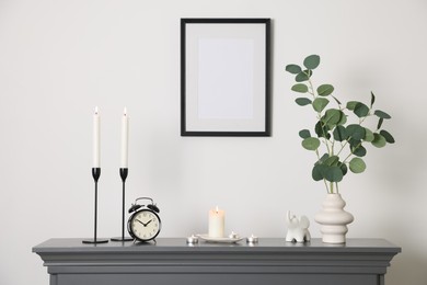 Photo of Stylish candlesticks with burning candles, other decor and alarm clock on fireplace near white wall indoors