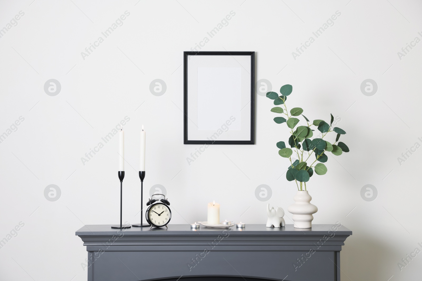 Stylish candlesticks with burning candles, other decor and alarm clock on fireplace near white wall indoors Photo of Stylish candlesticks with burning candles, other decor and alarm clock on fireplace near white wall indoors