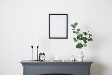 Photo of Stylish candlesticks with burning candles, other decor and alarm clock on fireplace near white wall indoors