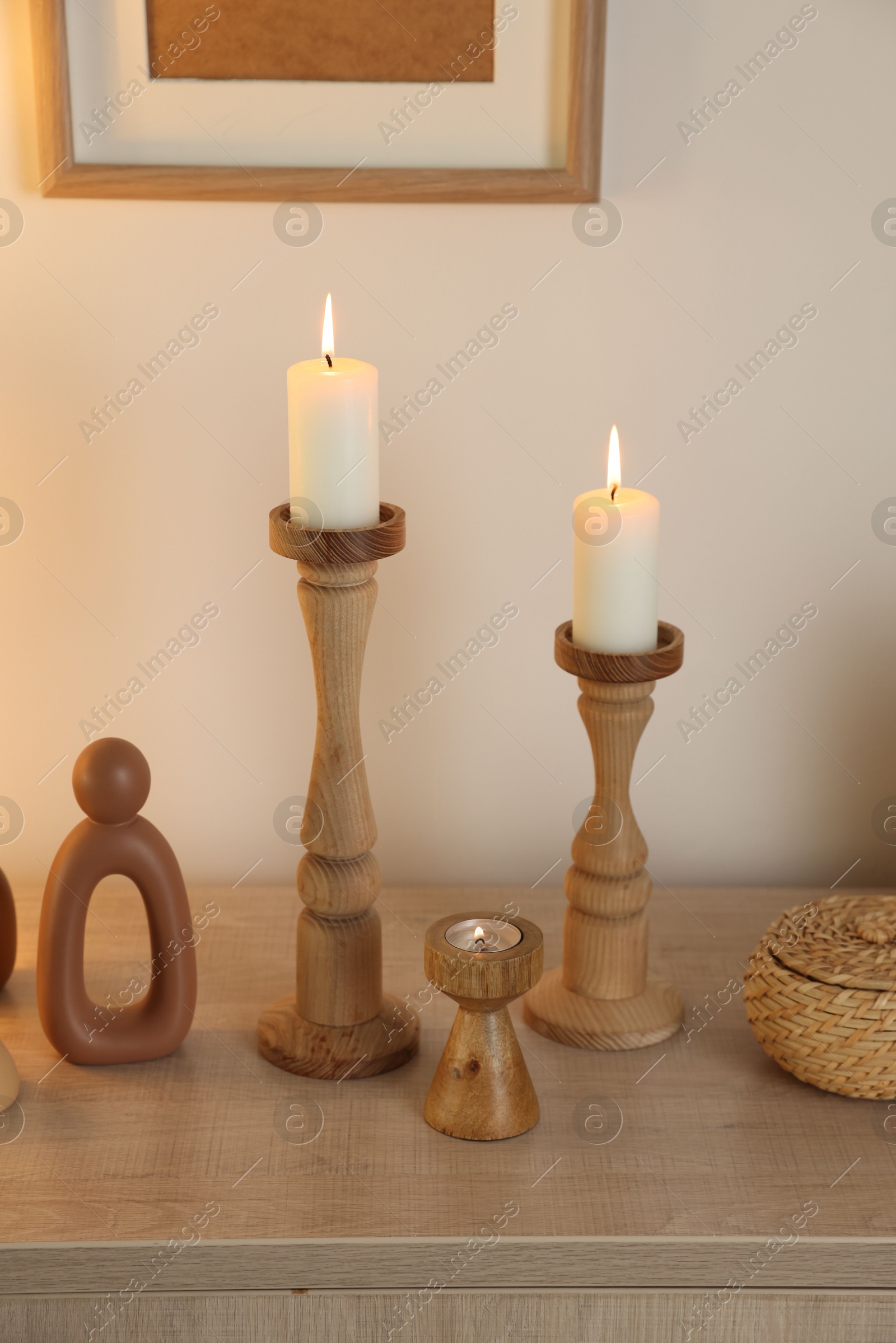 Stylish candlesticks with burning candles and other decor on cabinet near beige wall indoors Photo of Stylish candlesticks with burning candles and other decor on cabinet near beige wall indoors