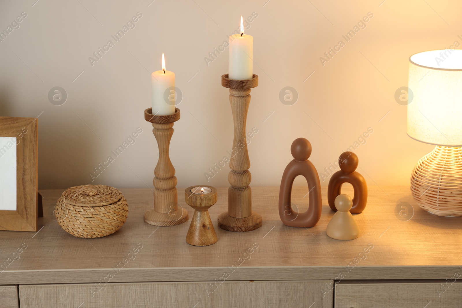 Stylish candlesticks with burning candles and other decor on cabinet near beige wall indoors Photo of Stylish candlesticks with burning candles and other decor on cabinet near beige wall indoors