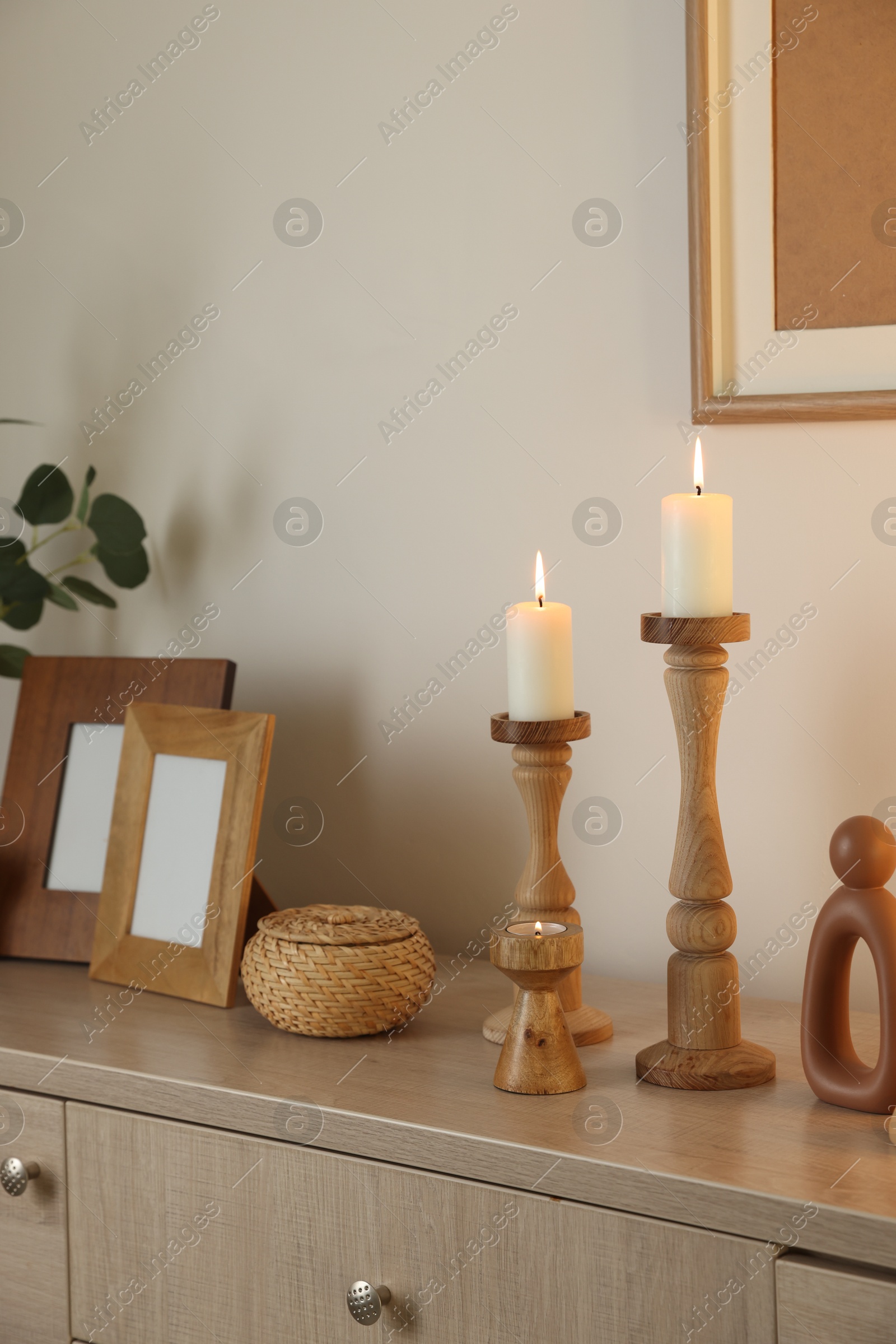 Stylish candlesticks with burning candles and other decor on cabinet near beige wall indoors Photo of Stylish candlesticks with burning candles and other decor on cabinet near beige wall indoors