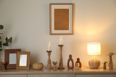 Photo of Stylish candlesticks with burning candles and other decor on cabinet near beige wall indoors