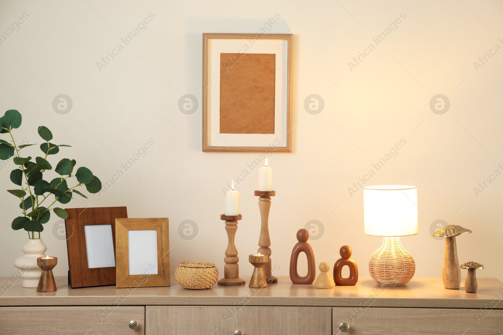 Stylish candlesticks with burning candles and other decor on cabinet near beige wall indoors Photo of Stylish candlesticks with burning candles and other decor on cabinet near beige wall indoors