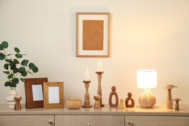 Photo of Stylish candlesticks with burning candles and other decor on cabinet near beige wall indoors