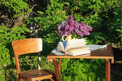 Photo of Beautiful lilac flowers in vase and book on wooden table in garden