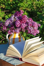 Photo of Beautiful lilac flowers in vase and book on table in garden, closeup