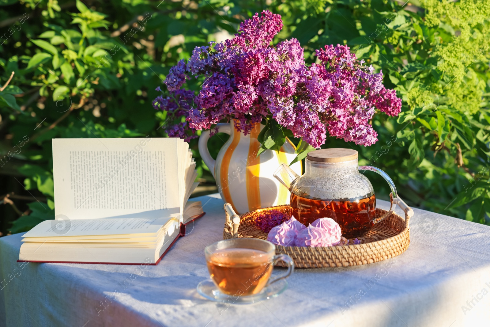 Beautiful lilac flowers in vase, tea, zephyrs and book on table in garden Photo of Beautiful lilac flowers in vase, tea, zephyrs and book on table in garden