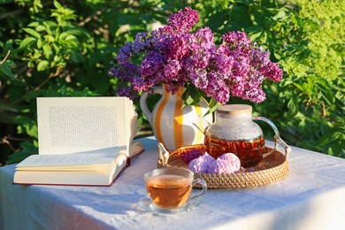 Photo of Beautiful lilac flowers in vase, tea, zephyrs and book on table in garden