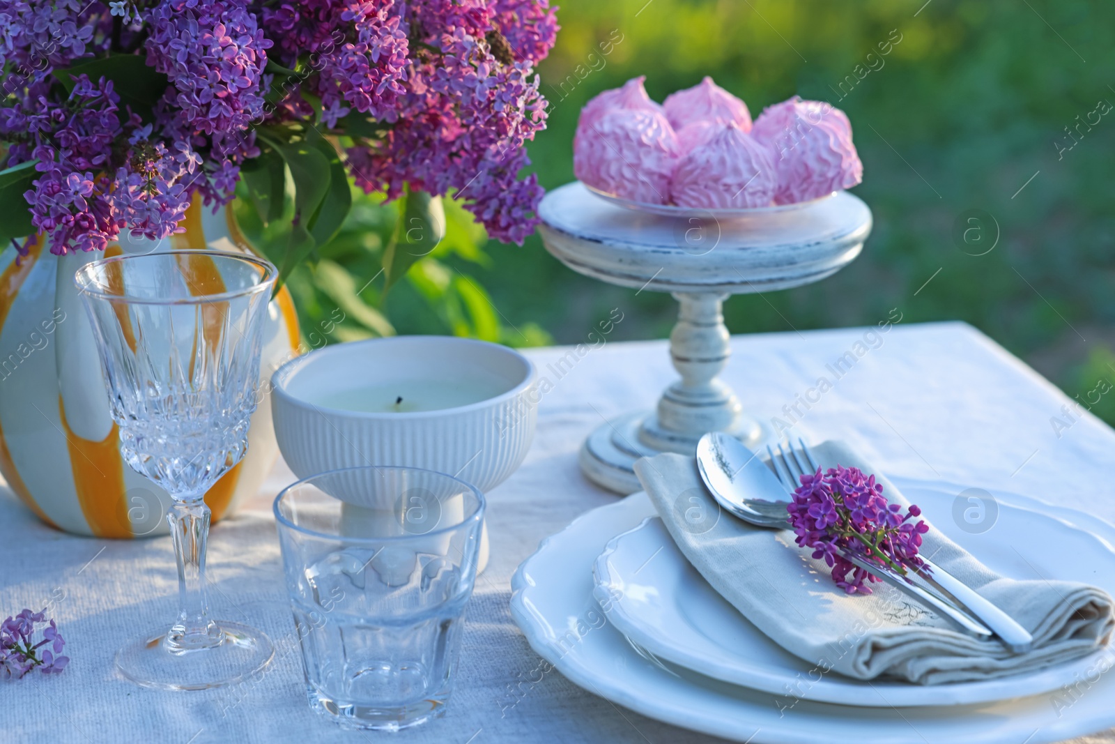 Beautiful lilac flowers, zephyrs, plates, cutlery, glasses and candle on table in garden, closeup Photo of Beautiful lilac flowers, zephyrs, plates, cutlery, glasses and candle on table in garden, closeup