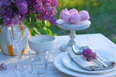 Photo of Beautiful lilac flowers, zephyrs, plates, cutlery, glasses and candle on table in garden, closeup