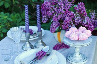 Photo of Beautiful lilac flowers, zephyrs, plates, cutlery, glasses and candles on table in garden, closeup