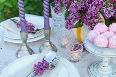 Photo of Beautiful lilac flowers, zephyrs, plates, cutlery, glass and candles on table in garden, closeup