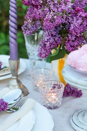 Photo of Beautiful lilac flowers, plates, cutlery and burning candles on table in garden, closeup