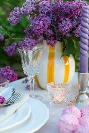 Photo of Beautiful lilac flowers, zephyrs, plates, cutlery, glass and candles on table in garden, closeup