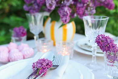 Photo of Beautiful lilac flowers, zephyrs, plates, cutlery, glasses and burning candles on table in garden, closeup