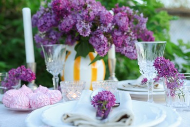 Photo of Beautiful lilac flowers, zephyrs, plates, cutlery, glasses and burning candles on table in garden, closeup