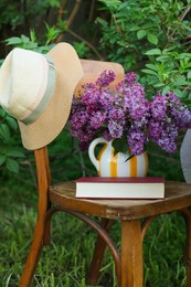 Photo of Beautiful lilac flowers in vase and book on wooden chair in garden