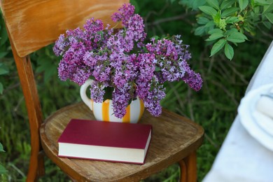 Photo of Beautiful lilac flowers in vase and book on wooden chair in garden