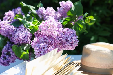 Photo of Beautiful lilac flowers, book and straw hat on table in garden, closeup