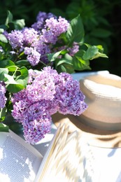 Photo of Beautiful lilac flowers, book and straw hat on table in garden, closeup