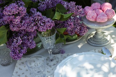 Photo of Beautiful lilac flowers, sweets, plates and glasses on table in garden, closeup