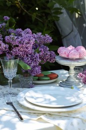 Photo of Beautiful lilac flowers, sweets, plates, glass and cutlery on table in garden, closeup