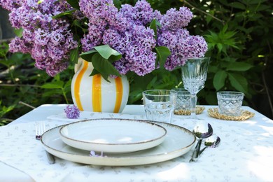 Photo of Beautiful lilac flowers, plates, cutlery, glasses and candles on table in garden, closeup
