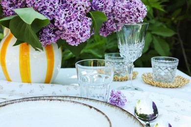 Photo of Beautiful lilac flowers, plates, spoon, glasses and candles on table in garden, closeup