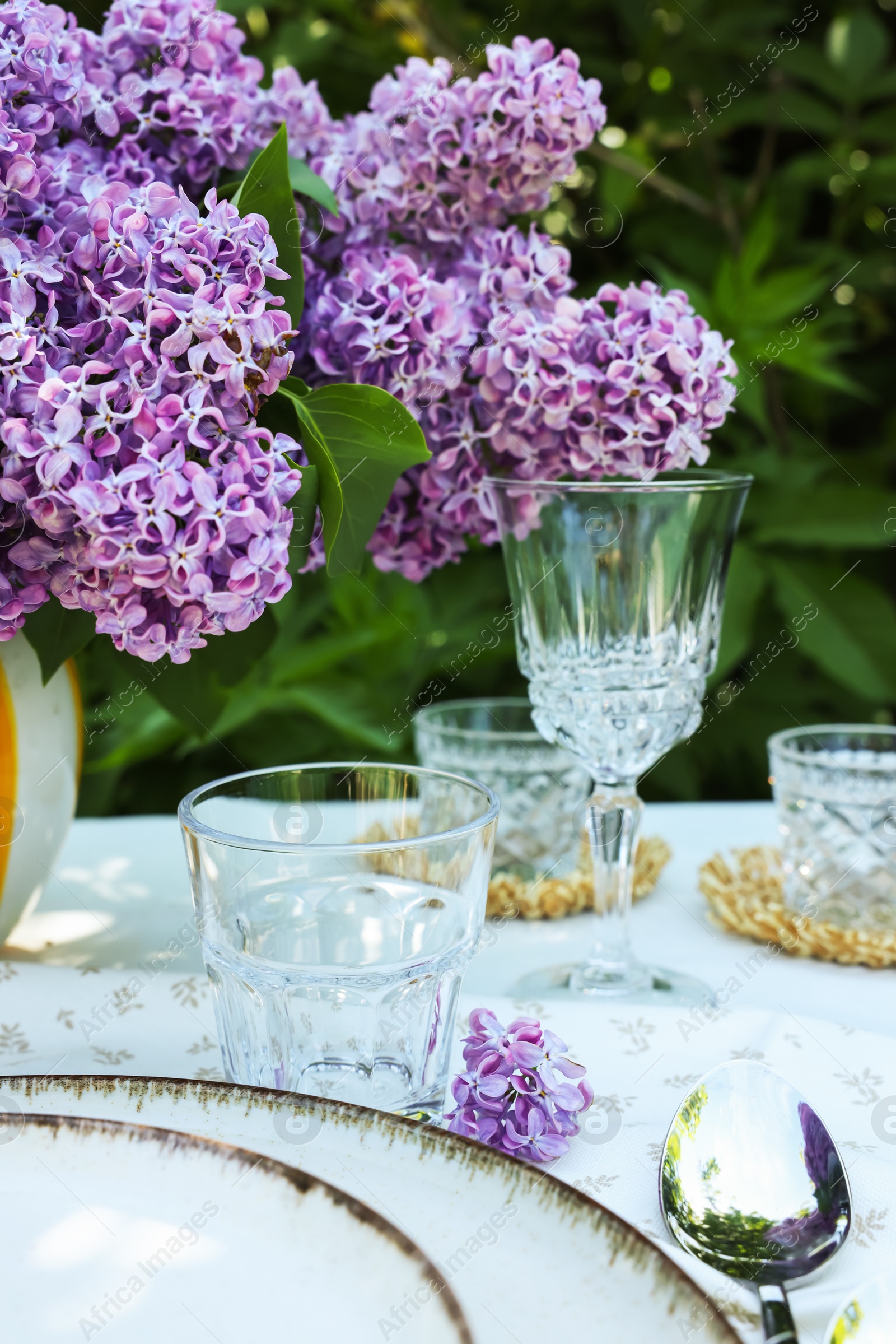 Beautiful lilac flowers, plates, spoon, glasses and candles on table in garden, closeup Photo of Beautiful lilac flowers, plates, spoon, glasses and candles on table in garden, closeup