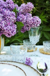 Photo of Beautiful lilac flowers, plates, spoon, glasses and candles on table in garden, closeup