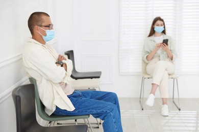 Photo of Man and woman in medical masks waiting for appointment indoors, selective focus