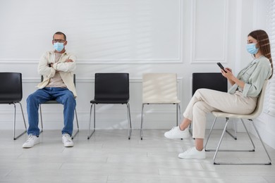 Photo of Man and woman in medical masks waiting for appointment indoors