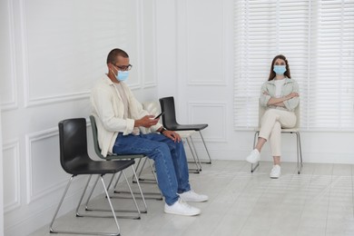 Photo of Man and woman in medical masks waiting for appointment indoors