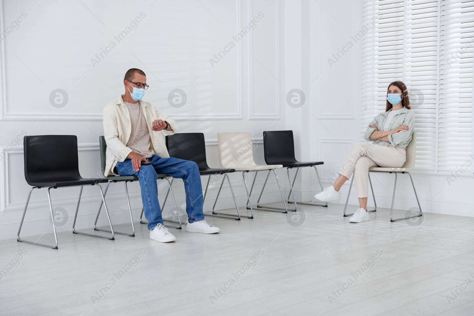 Man and woman in medical masks waiting for appointment indoors Photo of Man and woman in medical masks waiting for appointment indoors