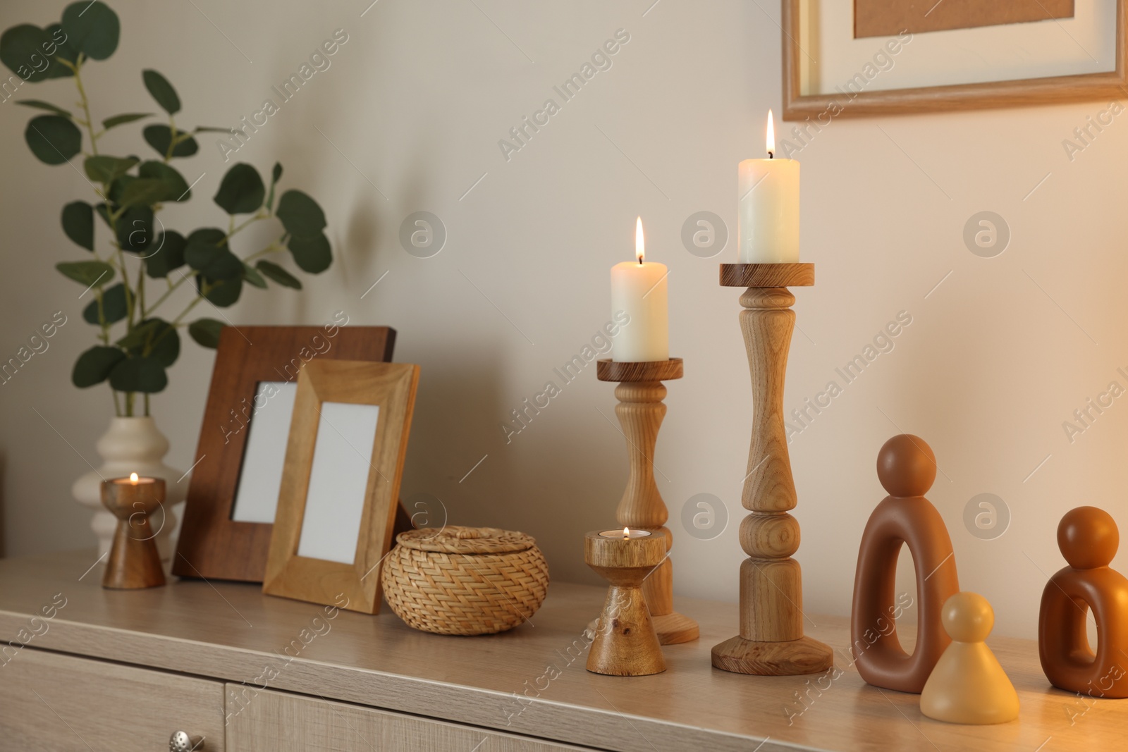 Stylish candlesticks with burning candles and other decor on cabinet near beige wall indoors Photo of Stylish candlesticks with burning candles and other decor on cabinet near beige wall indoors