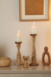 Photo of Stylish candlesticks with burning candles and other decor on cabinet near beige wall indoors