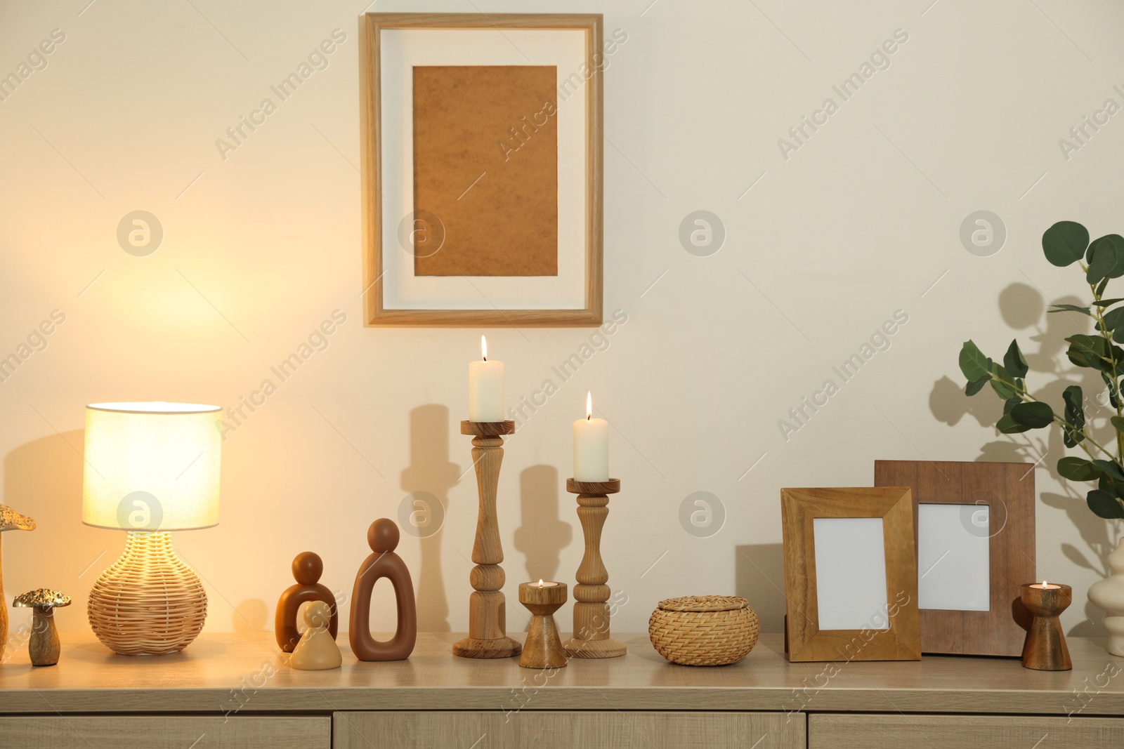Stylish candlesticks with burning candles and other decor on cabinet near beige wall indoors Photo of Stylish candlesticks with burning candles and other decor on cabinet near beige wall indoors